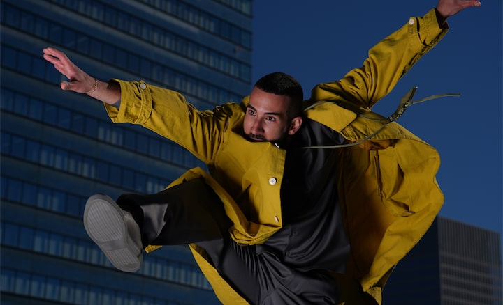 Devant un arrière-plan figurant des immeubles en ville, un homme aux cheveux courts qui porte une veste jaune est capturé en train de sauter.