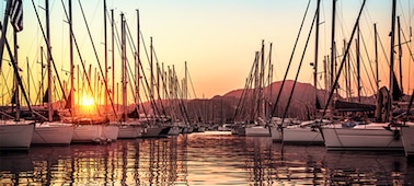 Sailing boats docked in bay with sunset reflected on the water and sun behind hills on the horizon