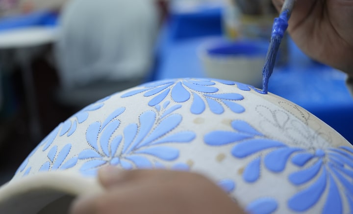 Exemple d’image – artisan ornant une jarre blanche de motifs bleus avec un pinceau. La mise au point est faite sur le pinceau. La jarre en premier plan et l’arrière-plan sont flous.